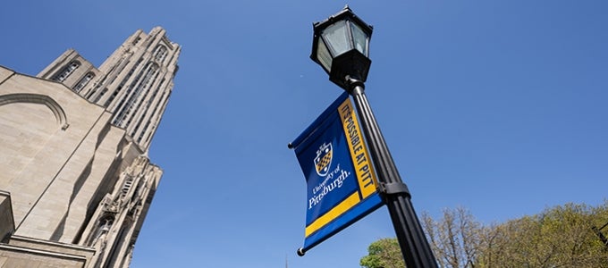 A blue-and-gold branded Pitt banner reading "It's Possible at Pitt" hangs on a lamp post with the Cathedral of Learning in the background. 