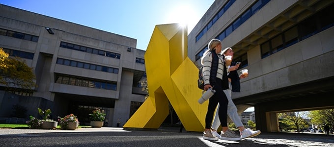 Two students walk by the yellow metal sculture "Light Up" on the Posvar patio.