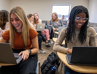 two smiling students working on laptops during class activity